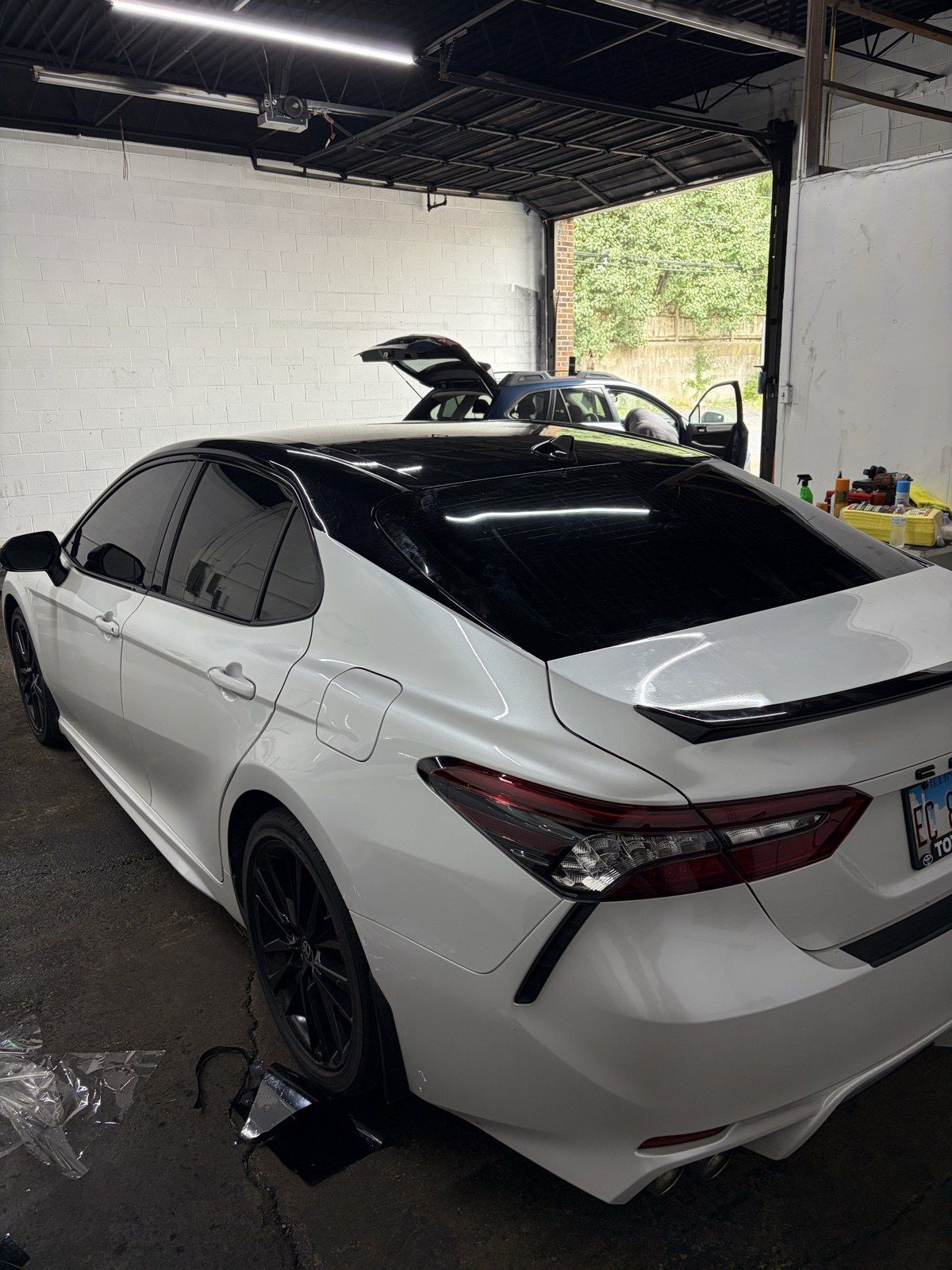 White Toyota Camry with a gloss-black roof wrap and tint, rear three-quarter view in the shop